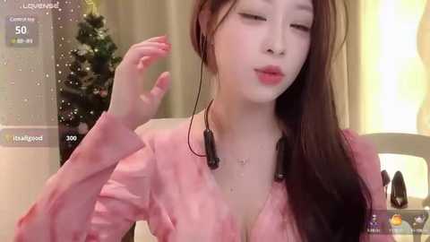 Media: Video of a fair-skinned East Asian woman with long brown hair, wearing a pink blouse, making a peace sign, and using headphones. Background includes a Christmas tree.