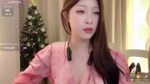 Media: Video of an East Asian woman with fair skin and long, straight brown hair, wearing a pink, lace-trimmed top, standing in front of a decorated Christmas tree and a lit room.