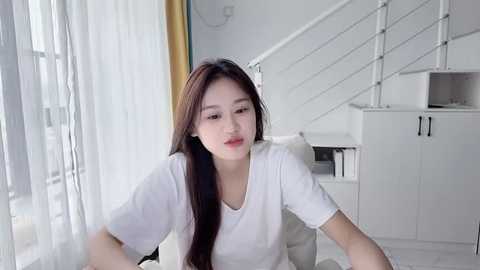 Media: Video of an Asian woman with long, straight brown hair, wearing a white t-shirt, sitting on a white chair in a modern, minimalist living room with white walls, stairs, and sheer white curtains.