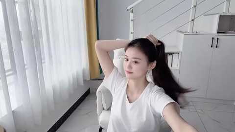 Media: Video of an Asian woman with long black hair in a white T-shirt, sitting on a white chair in a modern, minimalist room with white walls, glass doors, and a staircase.