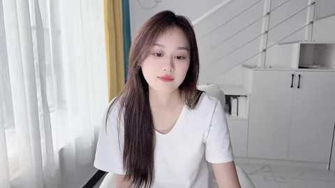 Media: A video of an Asian woman with long dark hair, fair skin, and a slender build, wearing a white t-shirt, standing indoors in a modern, minimalist room with white walls, a staircase, and white curtains.