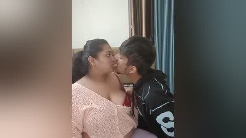 Media: A video captures an intimate moment between a young woman with medium brown skin, wearing a light pink off-shoulder top, and a young man with dark skin, wearing a black jacket, kissing passionately. The background shows a beige wall and blue curtains.