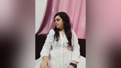 Media: Video of a woman with long black hair, wearing a white button-up shirt, sitting against a backdrop of pink and white fabric. Her expression is neutral, and she appears to be in a casual, indoor setting.