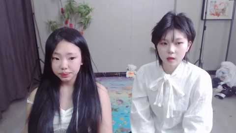 Media: Video of two East Asian women with long black hair, one wearing a white blouse, the other a white dress, sitting on a colorful rug in a sparsely decorated room.