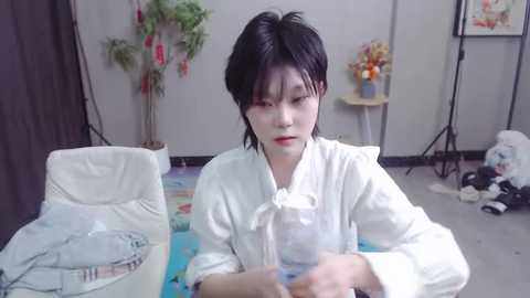 Media: Video of a young Asian person with short black hair, wearing a white robe, seated in a cluttered room with a white chair, plants, and a colorful rug.