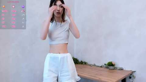 Media: A video of an Asian woman with straight black hair, wearing a white crop top and loose pants, adjusting her glasses in a minimalist room with a wooden table and potted plant.