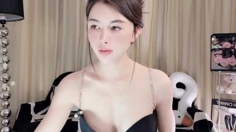 Media: Video of a young Asian woman with fair skin, brown hair, and slender build, wearing a black lace bra, sitting in a room with beige curtains and black lamp.