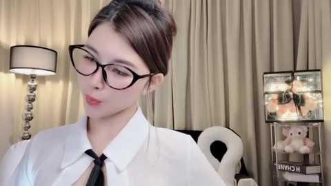 Media: A video of a fair-skinned woman with dark hair tied back, wearing black-rimmed glasses and a white shirt, standing in a room with beige curtains, a white lamp, and a \"MOM\" sign.