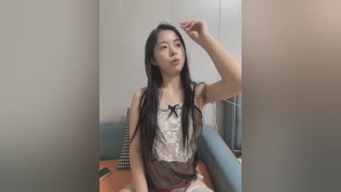 Media: A video of an Asian woman with long black hair, wearing a sheer lace top and red skirt, sitting on a blue couch in a minimalist room.