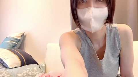 Media: Video of a young East Asian woman with shoulder-length brown hair, wearing a white surgical mask and a gray sleeveless top. She is in a bedroom with a bed featuring blue and white pillows.