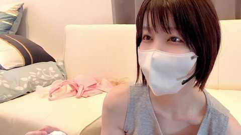 Media: Video of a young Asian woman with short, dark brown hair, wearing a white face mask, gray tank top, and black headset, seated on a cream couch in a cozy, minimally decorated room.