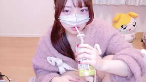 Media: Video of an East Asian woman with straight, brown hair, wearing a pink, fuzzy bathrobe, white face mask, and drinking from a plastic bottle. Background features a white wall and a plush toy.