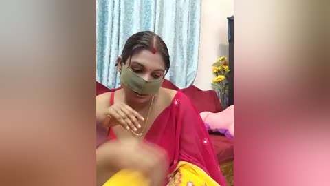 Media: Video of a South Asian woman in a red sari with a yellow border, wearing a green mask, seated on a red sofa, hands on her chest. Background includes light blue curtains, a vase of yellow flowers, and a pink cushion.