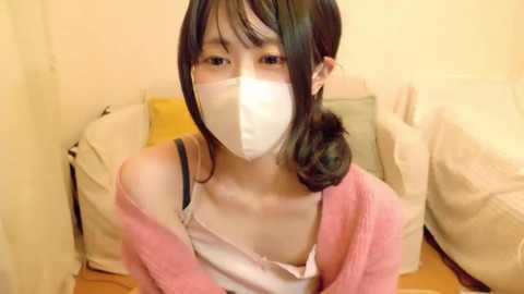 Media: Video of an Asian woman with straight black hair, wearing a pink cardigan, white mask, and a white top, sitting on a bed with beige and yellow pillows.