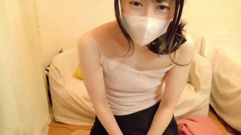 Media: Video of a young Asian woman with shoulder-length black hair, wearing a white tank top, black skirt, and a white surgical mask, seated in a dimly lit, cream-colored room.