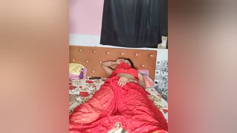 Media: Video of a woman in red saree, lying on a bed with floral sheets, head covered, in a pink and white room with a black curtain.