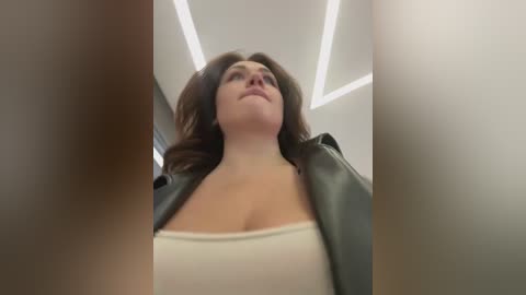 Media: A low-angle video of a woman with light skin, long brown hair, and a black leather jacket, wearing a white tank top, standing in a modern, brightly lit room with recessed lights.