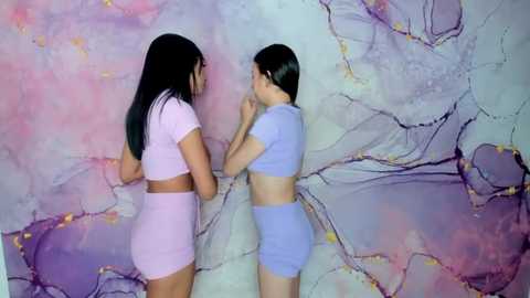 Media: Video of two young women with long black hair, wearing matching light pink and blue crop tops and shorts, painting a pastel marble mural on a wall.