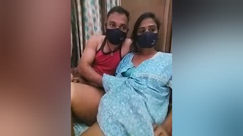 Media: Video of a man and woman in a hospital room, both wearing masks, with the woman in a blue hospital gown, sitting on a bed.