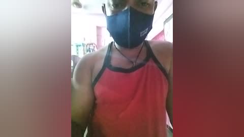 Media: Video of a person wearing a black mask, red tank top, and a black shirt, standing indoors with a blurred background.