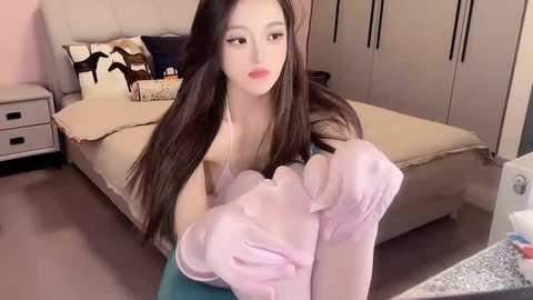 Media: Video of a young East Asian woman with long black hair, fair skin, and full lips, wearing pink gloves, sitting on a bed with beige linens and a white wall behind her.