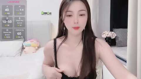 Media: A video of an Asian woman with long black hair, fair skin, and a slender figure, wearing a black lace bra, standing in a modern, minimalist bedroom with white walls and a bed.