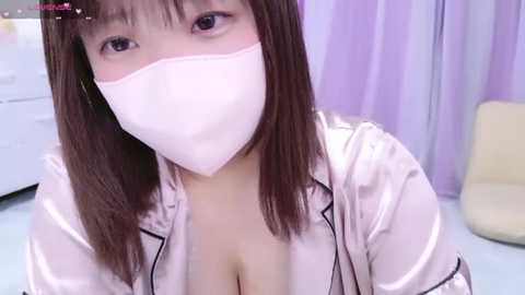 Media: A video of an East Asian woman with straight brown hair and a white surgical mask, wearing a pink satin robe with black trim, revealing cleavage, in a pastel-colored room with a white dresser and yellow chair.