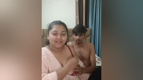 Media: Video of a young South Asian woman with medium skin tone, wearing a pink, textured robe, seated on a bed. Behind her, a young man with dark skin and short hair, shirtless, sits close. Background features a beige headboard and blue curtains.