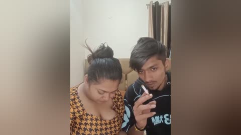 Media: A video of a young South Asian couple, both with medium skin tones, sitting closely. The woman, with a bun, wears a patterned top; the man, with tousled hair, holds a phone.