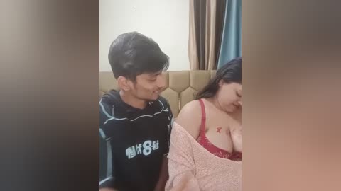 Media: Video of a young South Asian man in a black sports jersey and a woman with long dark hair and a red bra, sitting on a beige cushioned seat in a dimly lit room.