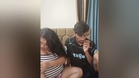 Media: Video of a young couple sitting on a beige couch. The man, with short dark hair, wears a black T-shirt, holding a phone. The woman, with long dark hair, wears a striped shirt, and is hugging the man. Background includes beige and blue curtains.