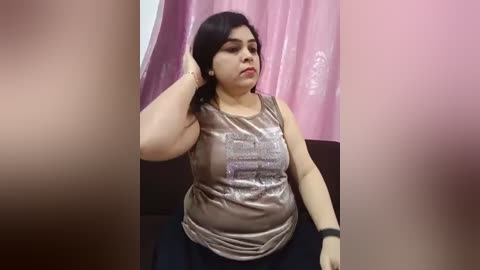 Media: Video of a plus-sized woman with medium skin tone and dark hair, wearing a glittery silver tank top, sitting in a chair against a pink curtain background.