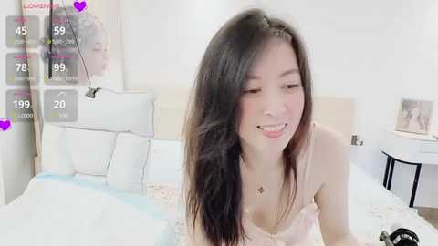 Media: A video of a young Asian woman with long black hair, wearing a pink camisole, smiling on a bed. The background features a minimalist bedroom with white walls, a headboard, and a bedside table.