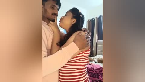 Media: Video of an intimate moment between a South Asian man and woman in a bedroom, wearing red and white striped dress, embracing passionately.