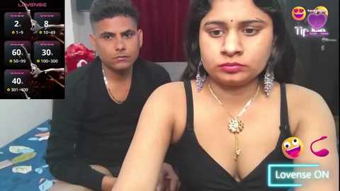 Media: Video of a South Asian woman with medium skin tone and black hair, wearing a black dress, and a man with short black hair, both sitting on a bed with a blue sheet.