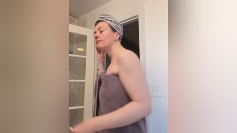 Media: Video of a fair-skinned, short-haired woman with a checkered headband, wrapped in a towel, standing in a bathroom with beige walls and white cabinets.