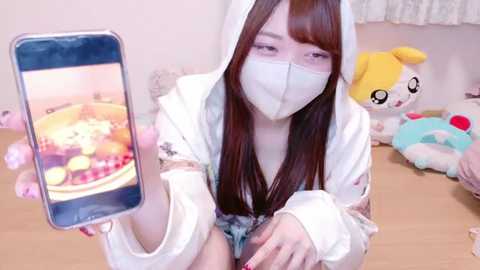 Media: Video of an Asian woman with long brown hair, wearing a white hooded sweatshirt and face mask, taking a selfie with a smartphone. Background includes stuffed animals, including a yellow bear and a blue dog.