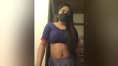 Media: Video of a young South Asian woman with medium skin tone, wearing a purple cropped top, blue pants, and a black face mask, standing indoors.