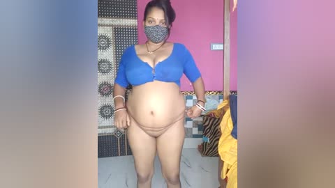 Media: Video of a pregnant South Asian woman with medium skin tone, wearing a blue crop top and brown panties, standing in a colorful room with a leopard-print rug, pink wall, and zebra-print curtains.