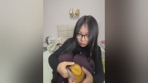 Media: A video of an Asian woman with long black hair, wearing glasses, and a black sweater, holding a yellow banana, in a messy bedroom.