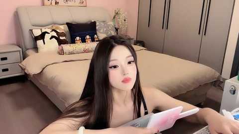 Media: A video of an East Asian woman with long black hair, wearing pink lipstick, lying on a beige bed in a modern bedroom, reading a book.