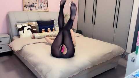 Media: Video of a woman lying on her back on a beige bed, wearing black pantyhose, her legs raised and spread, revealing her pink underwear. The room has light pink walls, a white headboard, and white wardrobes.