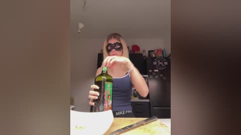 Media: Video of a woman with a black mask, blonde hair, and blue tank top pouring olive oil from a green bottle into a white bowl. Kitchen background with black appliances and red objects.