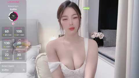 Media: Video of an Asian woman with fair skin, long black hair, and large breasts, wearing a white off-shoulder top, sitting on a bed in a brightly lit bedroom.