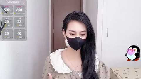 Media: Video of an East Asian woman with long black hair and fair skin, wearing a black face mask and beige dress with white lace trim, standing in a bathroom with a thermometer and temperature chart on the wall.