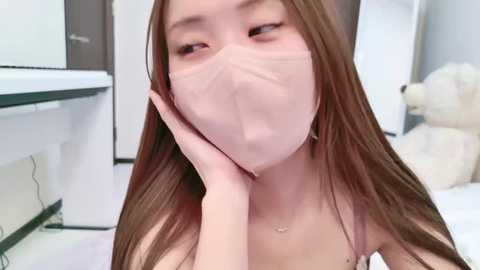 Media: Video of a young Asian woman with long brown hair, wearing a face mask, resting her chin on her hand, in a bright, modern room with white and gray furniture and a teddy bear.