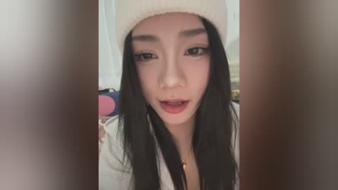 Media: A video of a young East Asian woman with long black hair, fair skin, and a light beige knit beanie, wearing a white shirt, smiling softly.