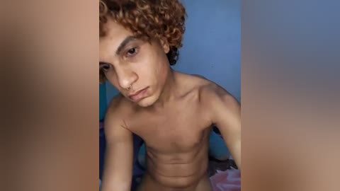 Video of a young, light-skinned, shirtless man with curly, reddish-brown hair, sitting indoors against a blue wall, looking slightly to the left.