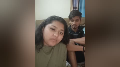 Media: A video of an Indian woman with medium skin tone and dark hair, wearing a green top, making a pouty face. Behind her, a man with medium skin tone and short hair, dressed in a black shirt, looks serious.