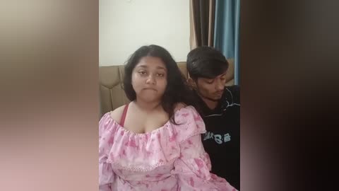 Media: Video of two South Asian women, one in a pink floral off-shoulder dress, the other in a black t-shirt, sitting side by side on a beige couch in a room with blue curtains and a light-colored wall.